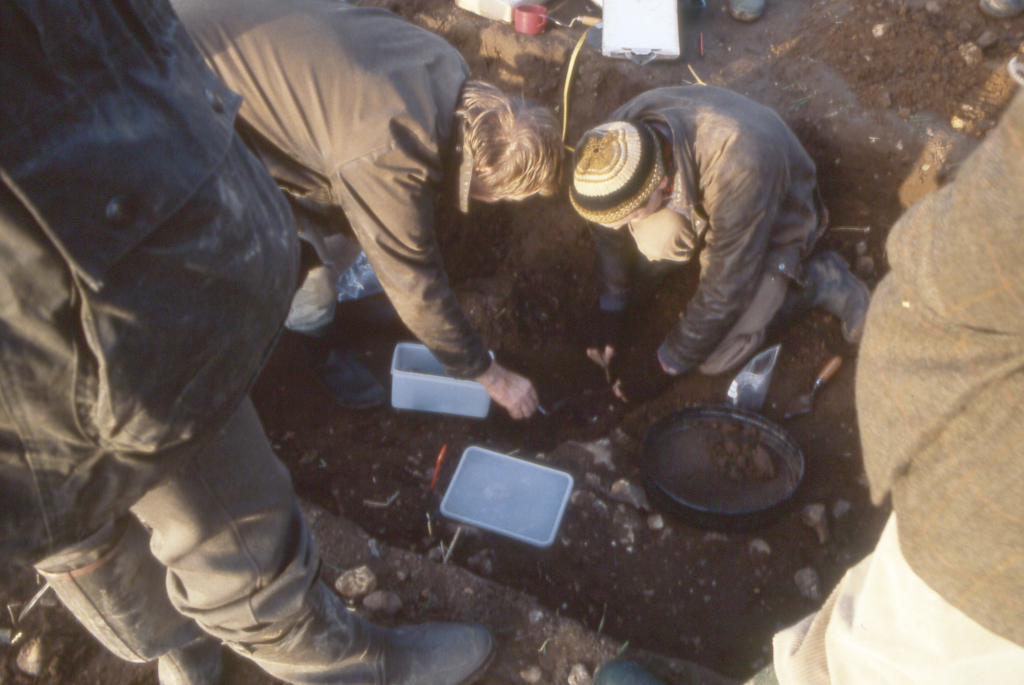 two archaeologists in exposed trench over various items to be recovered